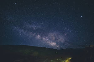 銀河系在黑暗山脈和燈火通明的村莊上方的星空夜空中延伸。 — 藍色銀河 壁紙