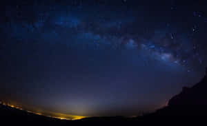 銀河系在黑暗山丘和遠處城市燈光之上的星空夜空中延伸。 — 星空動畫 壁紙