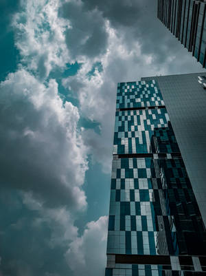 在戲劇性的多雲天空下，高聳的玻璃摩天大樓帶有格紋面板。 — 蒸氣波 壁紙