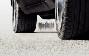 碎石上的汽車輪胎，後面有模糊的人走過。 — 玩命關頭 壁紙