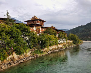 不丹寺廟位於河邊，被綠樹和山巒環繞，天空多雲 — 不丹 壁紙