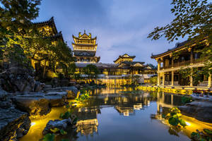 古老的中式亭閣在黃昏時亮起，倒映在寧靜的池塘中，周圍有岩石和樹木。 — 唐人街 壁紙