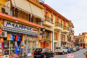色彩繽紛的商店和停放的汽車排列在一條陽光普照的地中海街道上，帶有陽台。 — 汽車 壁紙