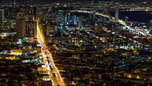繁華城市的夜間鳥瞰圖，有燈火通明的建築物和一座橫跨水面的燈光橋樑。 — 舊金山 壁紙