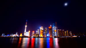 夜景，發光的城市天際線倒映在水中，上方有月亮。 — 唐人街 壁紙