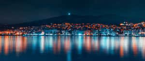 夜晚的城市燈光映照在平靜的水面上，背景有山脈。 — 3440x1440 壁紙