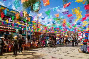 色彩繽紛的 papel picado 橫幅懸掛在熱鬧的戶外市場街道上，有棕櫚樹和購物者。 — 布宜諾斯艾利斯 壁紙