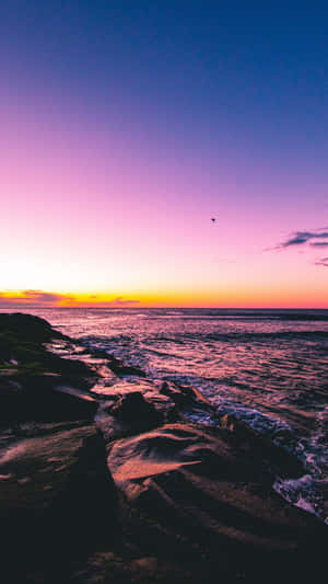 海洋上的日落，海浪在紫色橙色天空下拍打黑暗岩石 — 海灘 iPhone 壁紙
