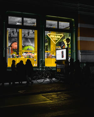夜晚在燈光明亮的 Prada 商店外的人和自行車剪影。 — Prada 壁紙