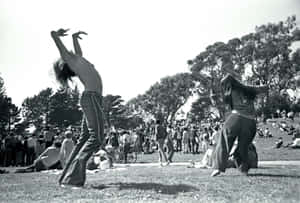 兩名舞者在陽光明媚的公園中，有樹木和圍觀的人群。 — Woodstock 壁紙