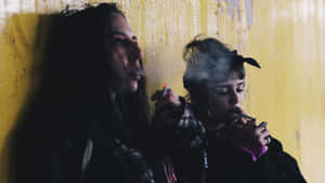 兩位女性靠著黃色牆壁，在陰暗的燈光下抽煙。 — 抽菸 壁紙