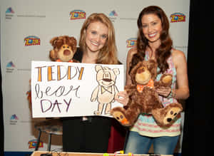 兩位女性在醫院活動中抱著泰迪熊和一個「Teddy Bear Day」標語牌。 — 安妮與艾比 壁紙