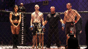 兩位拳手和裁判在比賽中站在有獎盃的MMA籠子裡。 — Pro Display XDR 壁紙