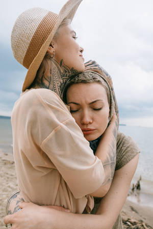 兩位紋身女士在海灘上擁抱，身後是海浪。 — 情侶擁抱 壁紙