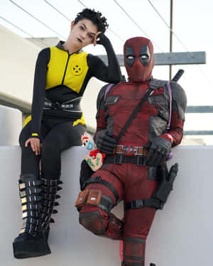 兩位穿著Deadpool和Jean Grey角色扮演服裝的人坐在戶外的白色邊緣上。 — 死侍 壁紙