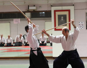 兩名男子在武術道場練習劍術，背景有學生坐著。 — 運動 壁紙