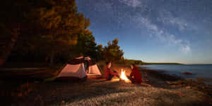 夜晚，星空下，兩人坐在湖邊的營火旁，旁邊有帳篷。 — 午夜 壁紙