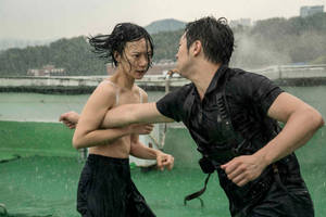 兩個人在雨中在屋頂上打架，身後是城市建築。