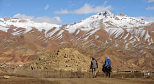 兩個人帶著一匹馬穿過一個有雪峰的山區沙漠景觀。 — 阿富汗 壁紙