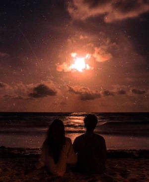 兩個人夜晚坐在沙灘上，透過雲層觀看月亮。 — 日落 壁紙