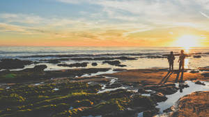 兩個人在日落時站在岩石海灘上觀看海洋 — 日落 壁紙