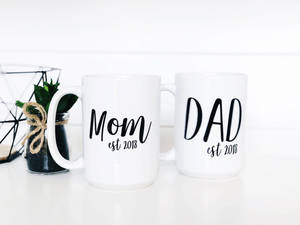 兩個白色馬克杯，上面寫著 Mom and Dad est 2018，旁邊有一棵小植物。 — 摩登家庭 壁紙