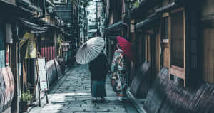兩個穿著傳統日本服裝的人，沿著一條有木建築的狹窄石巷走著。 — 夢幻 GIF 壁紙