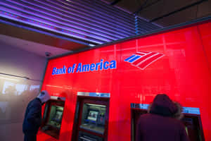 兩個在紅色牆壁和紫色天花板燈光的Bank of America分行內使用自動櫃員機的人。