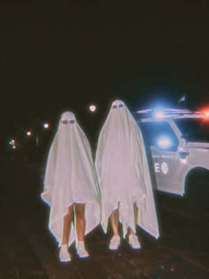 兩個裝扮成鬼魂的人夜間站在警車旁。 — 復古90年代風格 壁紙