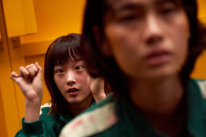 兩個穿著綠色制服的女孩在淺景深的橙色走廊中顯得很緊張。 — 067 魷魚遊戲 壁紙