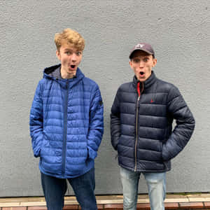 兩個穿著羽絨外套的年輕男子靠著灰色牆壁，震驚地睜大眼睛。 — 湯米英尼特 壁紙