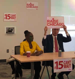 兩人在桌子旁舉著支持 raise chicago 和 15 cant wait 的抗議標語牌。 — Galaxy Z Flip7 FE 壁紙