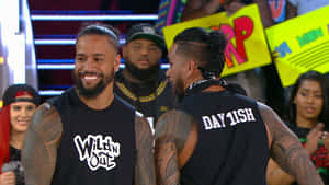 兩名微笑的紋身男子身穿黑色背心在舞台上，粉絲舉著標語牌。 — WWE GIF 壁紙