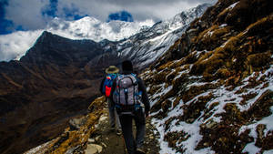 兩名遠足者正在攀登一條積雪的山徑，背景是崎嶇的山峰。 — 運動 壁紙