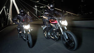 兩名騎士駕駛跑車在夜間燈火通明的橋上飛馳。 — 機車 壁紙