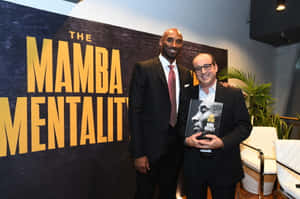 兩名身穿西裝的男子在活動中手持名為《The Mamba Mentality》的書籍擺姿勢。 — 科比· Bryant 壁紙