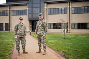 兩名身穿迷彩軍裝、面帶微笑的士兵站在建築物外的磚砌人行道上。 — MacBook Pro M3 壁紙