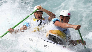 兩名皮划艇運動員在洶湧的白水中航行，表情專注，手持綠色槳。 — 奧運 壁紙
