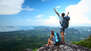 兩名背著背包的旅行者在懸崖邊緣歡呼，俯瞰熱帶景觀。 — 16:9 壁紙