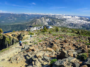 兩名遠足者在岩石山徑上，遠處有白雪覆蓋的山峰和森林。 — 登山 壁紙