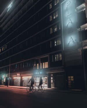 兩名騎自行車的人在夜間沿著城市街道騎行，經過一座有垂直招牌的明亮建築物。 — 7-ELEVEN 壁紙