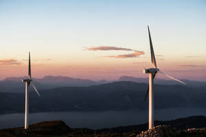 夕陽下，兩座風力發電機位於俯瞰山巒和海洋的山丘上。 — 能量 壁紙