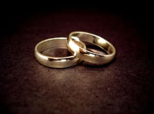 兩枚金色結婚戒指交纏在深色紋理表面上。 — 結婚戒指 壁紙