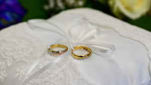 兩枚結婚戒指在白色蕾絲枕頭上，帶有花卉背景。 — 情侶 壁紙
