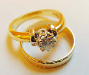 兩枚金結婚戒指，一枚鑲有單鑽，交纏在白色表面上 — 明亮 壁紙