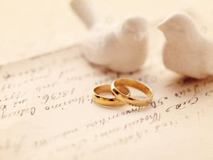 兩枚金色結婚戒指放在舊手寫信上，旁邊有白色小鳥雕像。 — 結婚戒指 壁紙