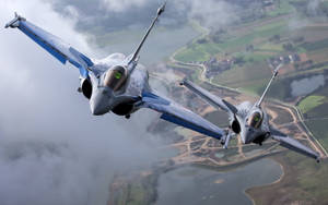 兩架戰鬥機飛越鄉村景觀，下方有田野和河流。 — 軍事動圖 壁紙