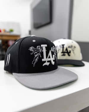 兩款以 LA 為主題、帶有天使設計的 snapback 帽展示在白色架子上。 — 4:5 壁紙
