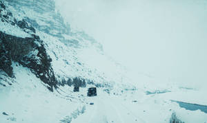 兩輛卡車在結冰河流旁的積雪山路上行駛。 — 山景 壁紙