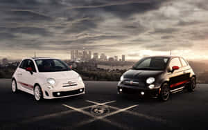 兩輛Fiat 500汽車停在瀝青路上，背景是夕陽下的城市天際線。 — Steam Deck 壁紙
